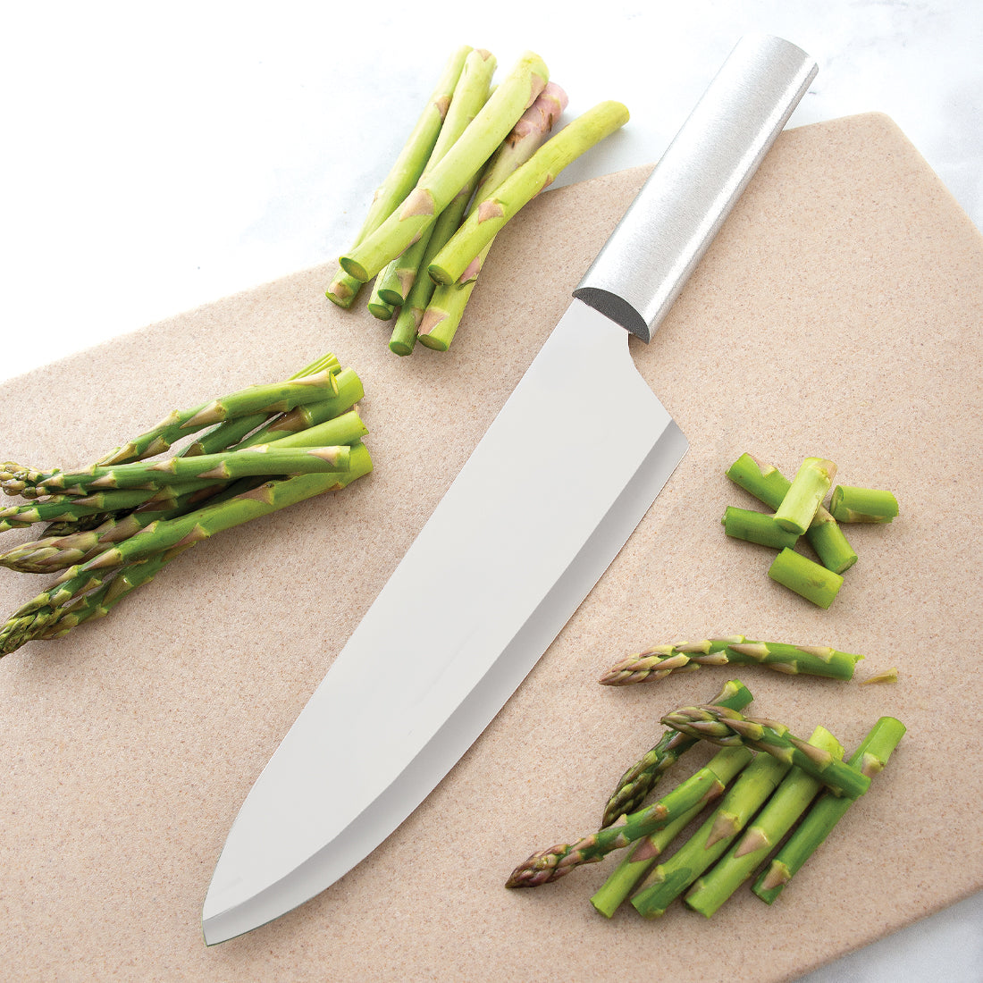 Handcrafted chef's knife with a long stainless steel blade and smooth metal handle, slicing fresh asparagus on a cutting board, part of USA Made kitchen and food prep collections.