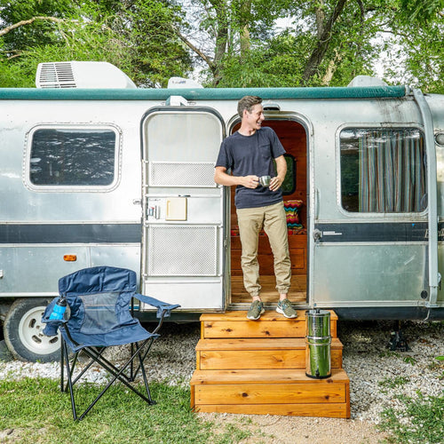 Man standing on wooden steps outside vintage camper trailer with stainless steel gravity water filter on steps and blue folding chair nearby in outdoor setting
