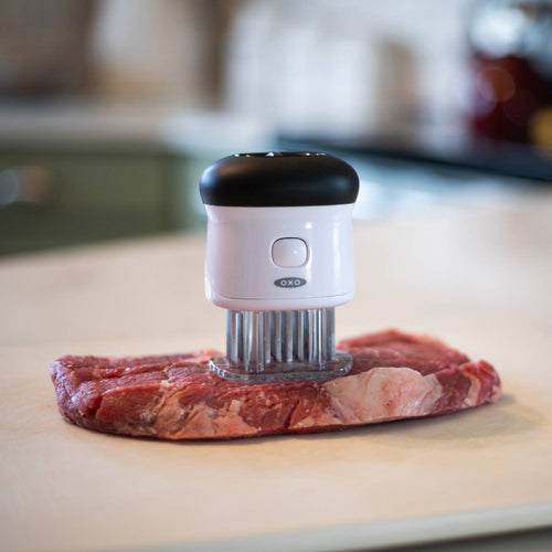 White and black meat tenderizer with 58 stainless steel blades pressing into a raw cut of beef on a countertop, featuring a safety lock and non-slip grip, ideal for Kitchen & Food, Specialty Equipment, Butchering Supplies, Best Sellers, and Promo Eligible collections.