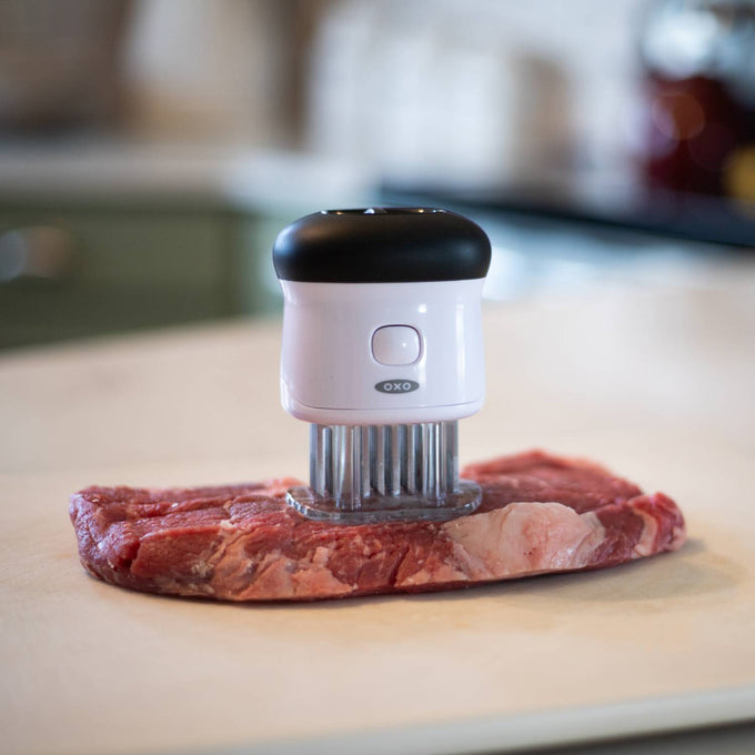 White and black meat tenderizer with 58 stainless steel blades pressing into a raw cut of beef on a countertop, featuring a safety lock and non-slip grip, ideal for Kitchen & Food, Specialty Equipment, Butchering Supplies, Best Sellers, and Promo Eligible collections.
