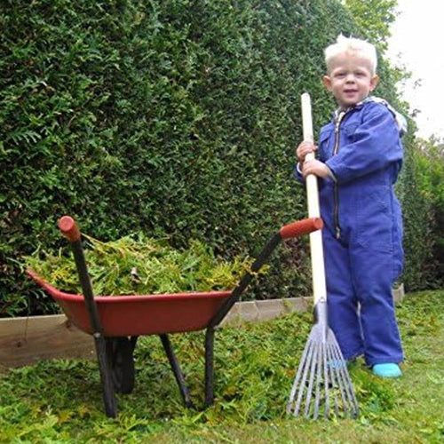 Young child in blue overalls holding a small rake next to a red wheelbarrow filled with green garden clippings, suitable for Farm, Garden & Tool and Lawncare collections.