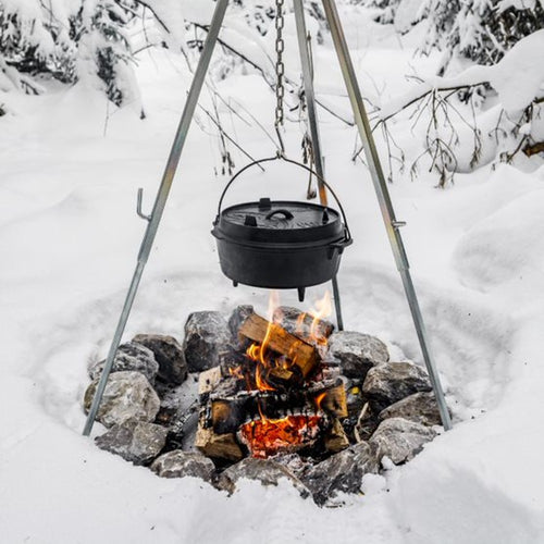 Black cast iron Dutch oven with legs hanging over an open fire pit surrounded by snow-covered rocks, showcasing outdoor cooking for Kitchen & Food, Cookware, Cast Iron collections.