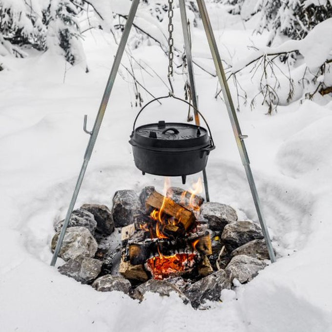 Black cast iron Dutch oven with legs hanging over an open fire pit surrounded by snow-covered rocks, showcasing outdoor cooking for Kitchen & Food, Cookware, Cast Iron collections.