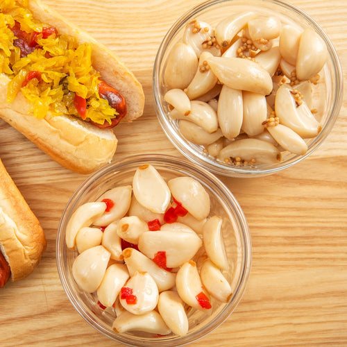 Two small glass bowls filled with sweet pickled garlic, one with mustard seeds and the other with bits of red chili, alongside a hot dog topped with relish on a light wooden surface, handcrafted and made in the USA, featured in Kitchen & Food, Food, Pantry, USA Made, Best Sellers, and Promo Eligible collections.