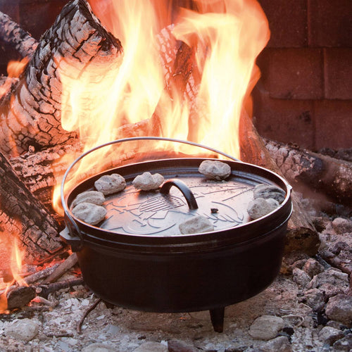 Seasoned cast iron camp Dutch oven with three legs and bail handle, cooking over campfire coals with dough pieces on the lid, handcrafted and made in USA, ideal for outdoor and camping cookware collections.
