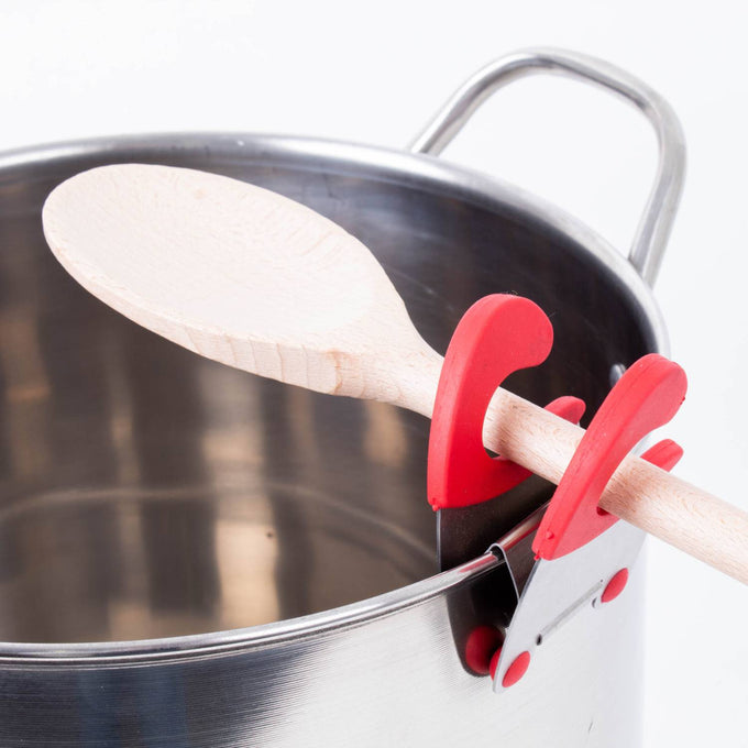 Red and silver pot clip spoon holder securely attached to a stainless steel pot, keeping a wooden spoon elevated and off surfaces, ideal for Kitchen Gadgets and Cooking Helpers.