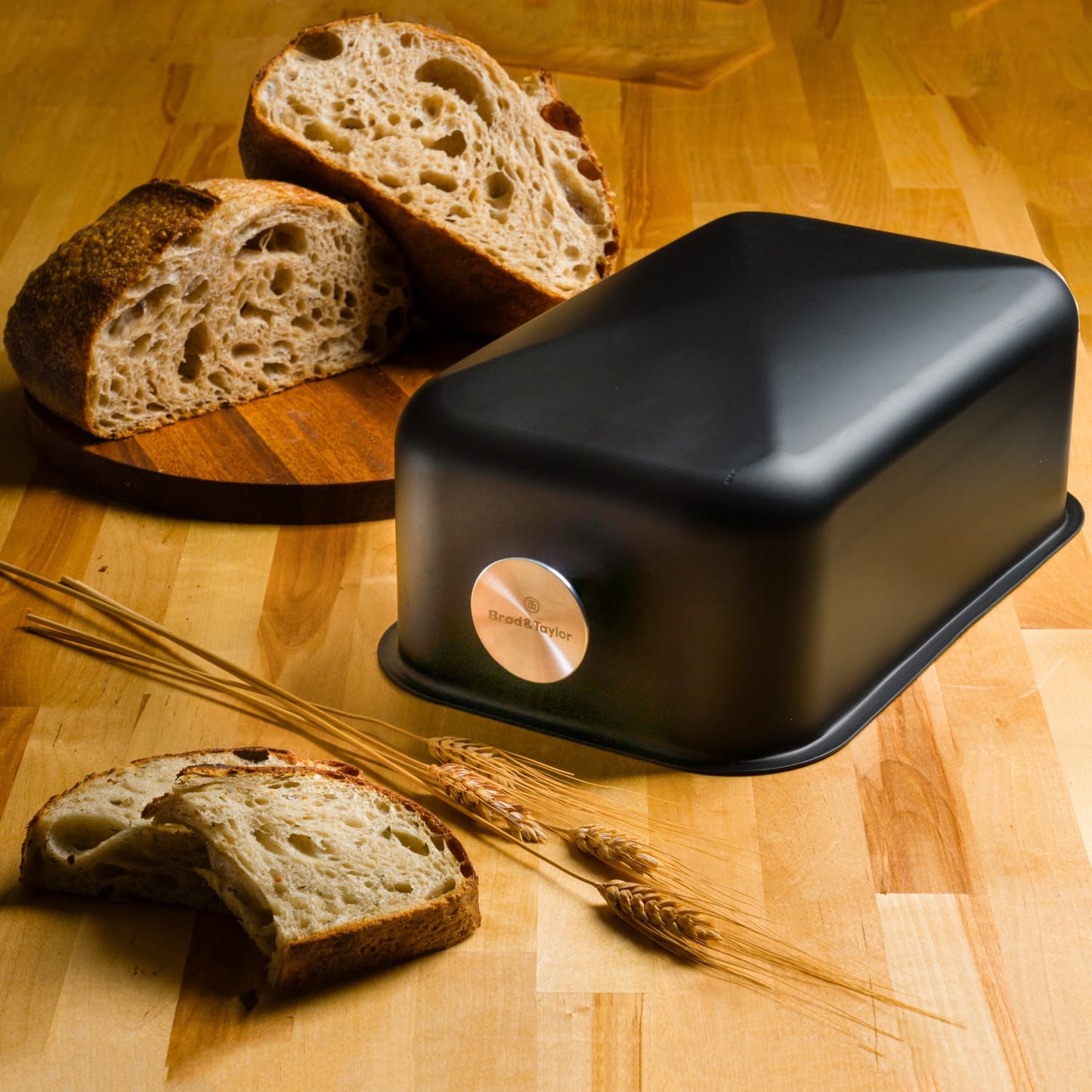 Matte black bread baking shell with smooth rounded edges and a silver logo medallion, shown with sliced artisan bread and wheat stalks on a wooden surface, ideal for Kitchen & Food and Baking Supplies collections.