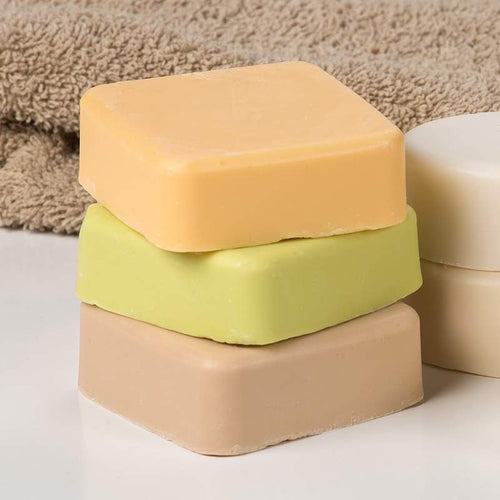 Three handcrafted square soap bars in muted tan, green, and orange colors stacked near a beige towel and white container, featured in Health & Wellness and Grooming collections.