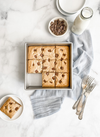 Square baking pan with freshly baked blondies cut into squares, placed on a light gray cloth alongside a bottle of milk, small bowl of chocolate pieces, and vintage forks, ideal for Kitchen & Food, Baking Supplies, and USA Made collections.