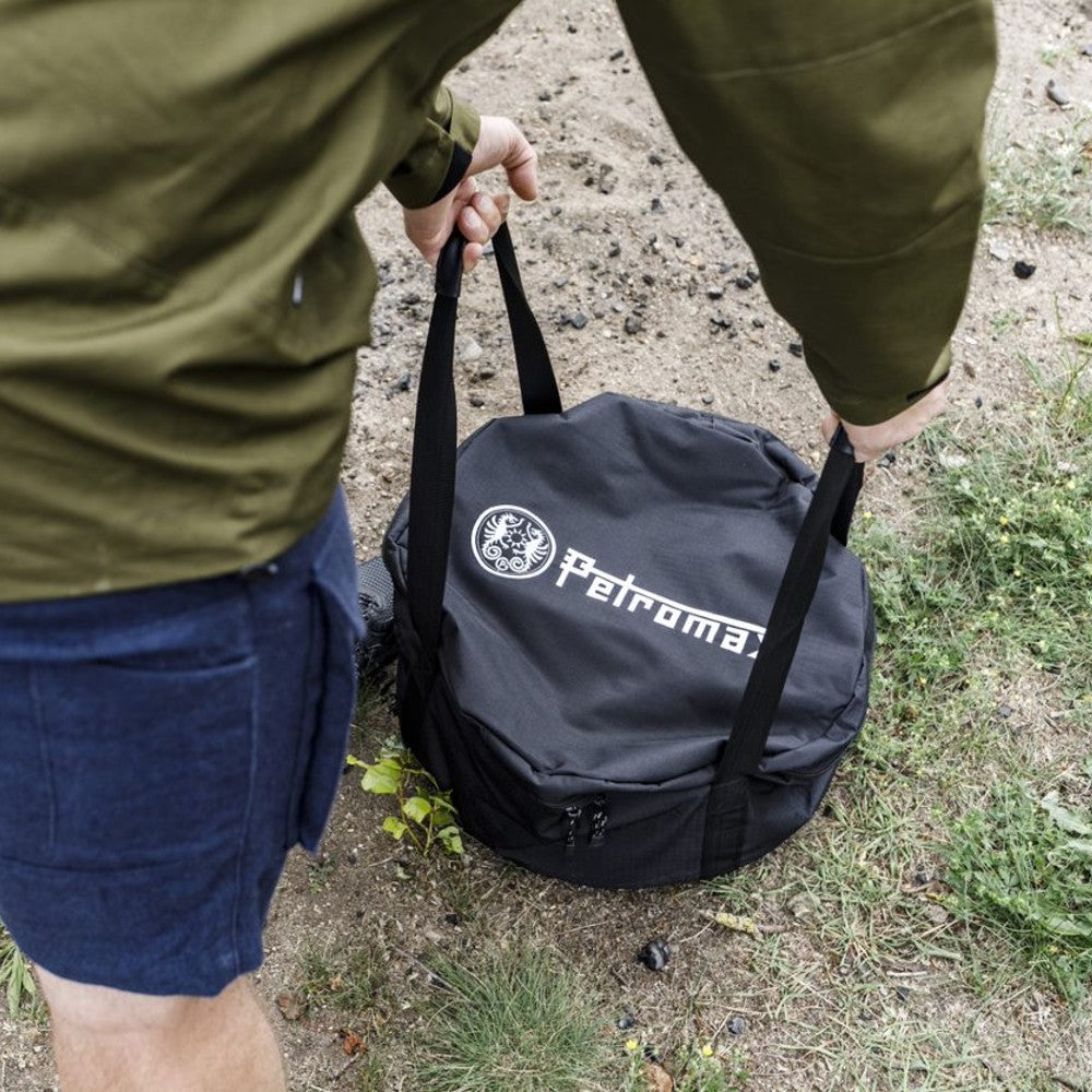Person carrying a black durable transport bag with Petromax logo, designed for safely storing cast iron cookware, shown outdoors on grass and dirt, suited for Farm, Garden & Tool, Camping, Best Sellers, and Promo Eligible collections.