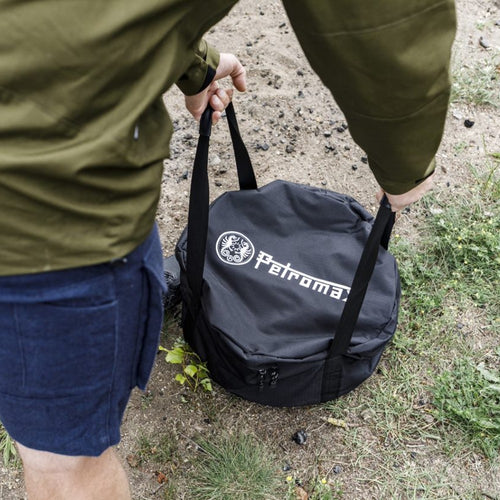 Person carrying a black durable transport bag with Petromax logo, designed for safely storing cast iron cookware, shown outdoors on grass and dirt, suited for Farm, Garden & Tool, Camping, Best Sellers, and Promo Eligible collections.