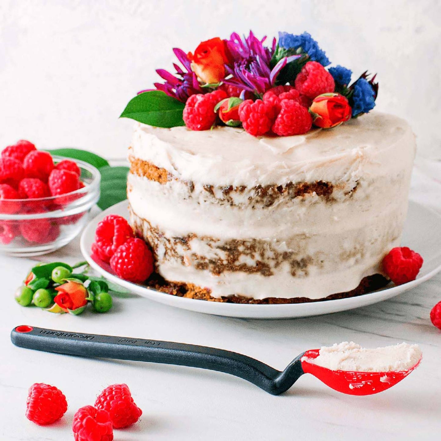 Flexible black and red silicone spoon with angled handle resting next to a frosted layer cake decorated with fresh raspberries and edible flowers, suited for Kitchen & Food, Food Prep, Measuring, and Cooking and Baking Helpers collections.