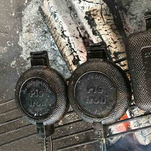 Two cast iron round pie irons with textured handles resting over hot campfire coals, showcasing durable cookware for camping and outdoor cooking collections.