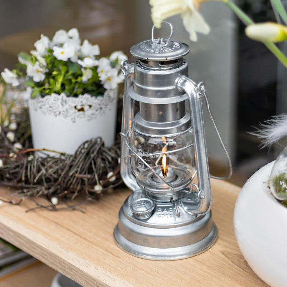 Handcrafted galvanized oil lantern with thermal heat-resistant globe emitting warm light, displayed on a wooden surface surrounded by floral decor, perfect for Lamps & Lighting, Camping, and Emergency Supplies collections.