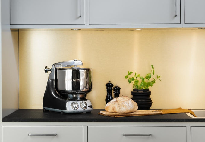 Ankarsrum Assistent Original Stand Mixer in black with stainless steel bowl on a kitchen countertop, accompanied by fresh bread, a potted herb, and salt and pepper shakers, ideal for Kitchen & Food, Baking Supplies, and Cooking and Baking Helpers collections.