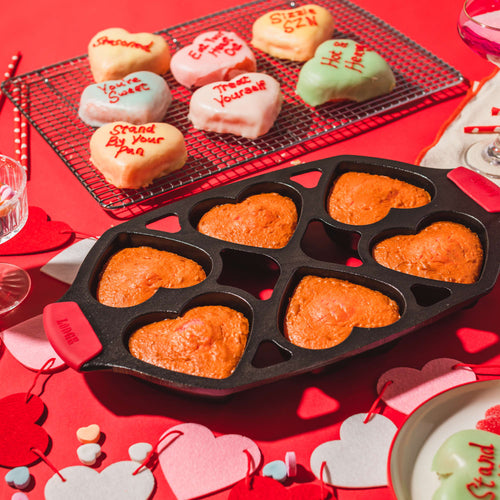 Handcrafted cast iron mini cake pan with six heart-shaped cavities filled with baked cakes, accompanied by decorated conversation heart cookies, featured in Kitchen & Food, Cast Iron, and USA Made collections.