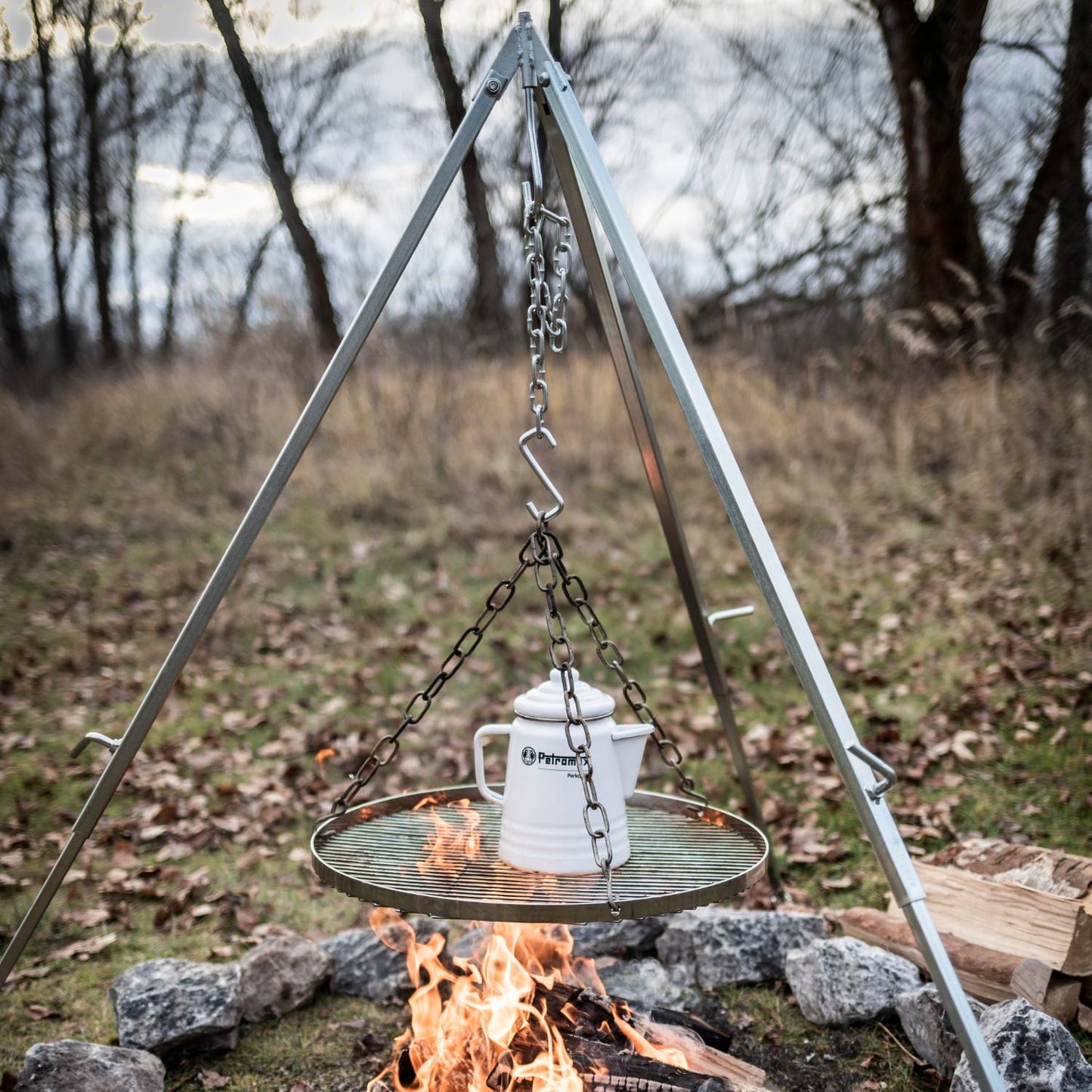 Chrome-plated steel hanging grate suspended by adjustable chains on a tripod over an open campfire, with a white pot on top, ideal for Farm, Garden & Tool, Camping, and Best Sellers collections.