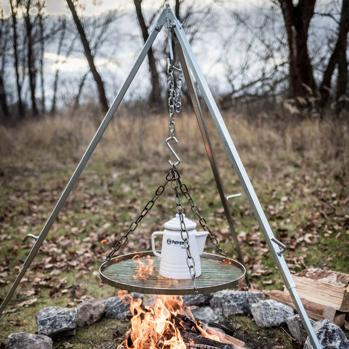 Chrome-plated steel hanging grate suspended by adjustable chains on a tripod over an open campfire, with a white pot on top, ideal for Farm, Garden & Tool, Camping, and Best Sellers collections.