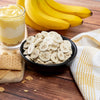 Black bowl filled with dried banana chips on a wooden surface, accompanied by fresh bananas, layered banana pudding dessert in a glass, graham crackers, and a yellow-accented kitchen towel, highlighting snack and pantry essentials in Farm, Garden & Tool, Emergency Supplies, Snacks and Sweets, USA Made, and Best Sellers collections.