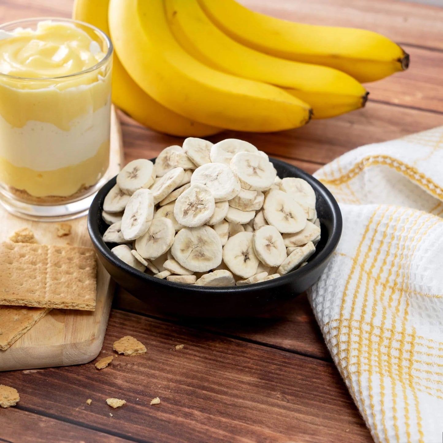 Black bowl filled with dried banana chips on a wooden surface, accompanied by fresh bananas, layered banana pudding dessert in a glass, graham crackers, and a yellow-accented kitchen towel, highlighting snack and pantry essentials in Farm, Garden & Tool, Emergency Supplies, Snacks and Sweets, USA Made, and Best Sellers collections.