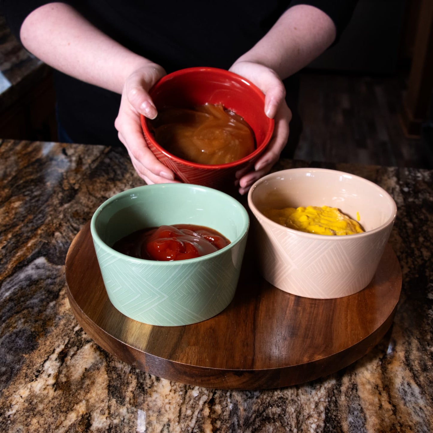 Three ceramic condiment bowls in red, green, and beige with subtle woven texture, filled with sauces and arranged on a handcrafted wooden Lazy Susan turntable in Kitchen & Food collections.