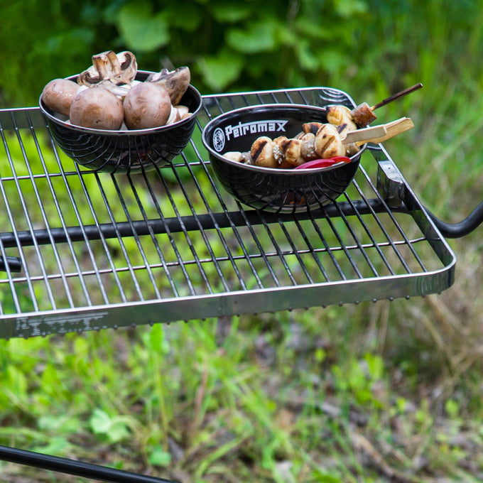 Two black enamel bowls with white logos, one holding mushrooms and the other grilled skewers, placed on a metal outdoor grill rack, suitable for Camping and Dinnerware collections.