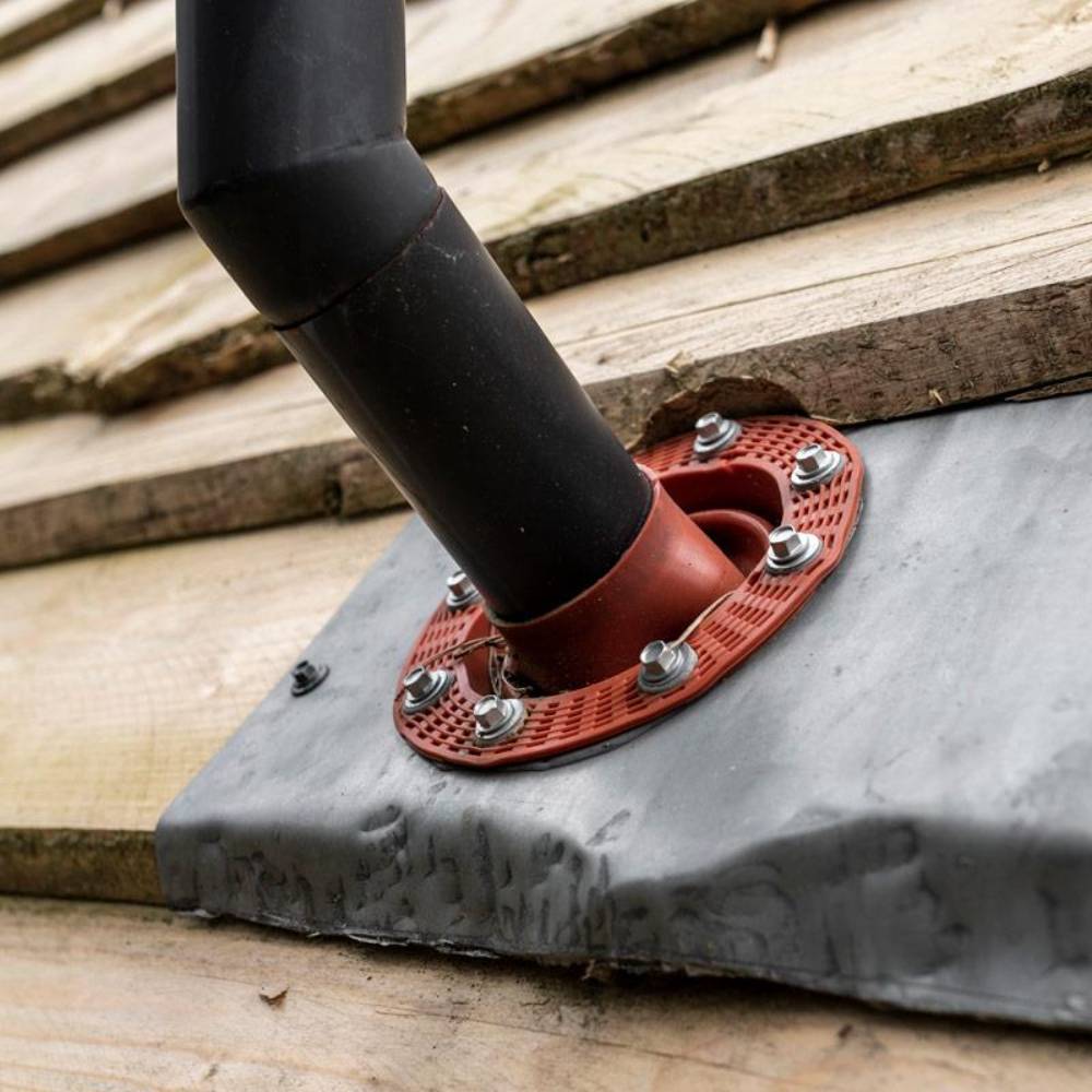 Close-up of a black metal pipe secured through a textured red flange with bolts, attached to weathered wooden planks and covered by a flexible protective metal flashing, suitable for Farm, Garden & Tool and Camping collections.