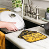 Two yellow and black scrub cloths with circular patterns, designed for gentle cleaning of cast iron and cookware, machine washable and made in USA, shown in a kitchen setting.