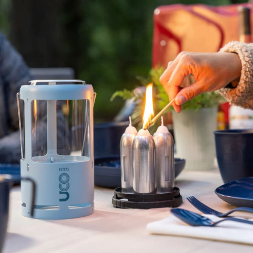 Handcrafted lightweight candle lantern with glass sides and metal frame, shown next to three tall candles being lit outdoors, ideal for camping, emergency supplies, and outdoor lighting.