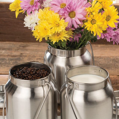 Set of three stainless steel milk cans with brushed finish, one holding fresh flowers, another filled with coffee beans, and the third containing milk, styled for rustic home decor and farm use.