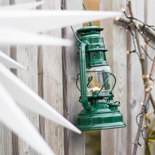 Handcrafted green galvanized oil lantern with thermal heat-resistant SUPRAX globe, glowing softly while hanging on a weathered wooden fence, perfect for lamps & lighting, camping, and emergency supplies collections.