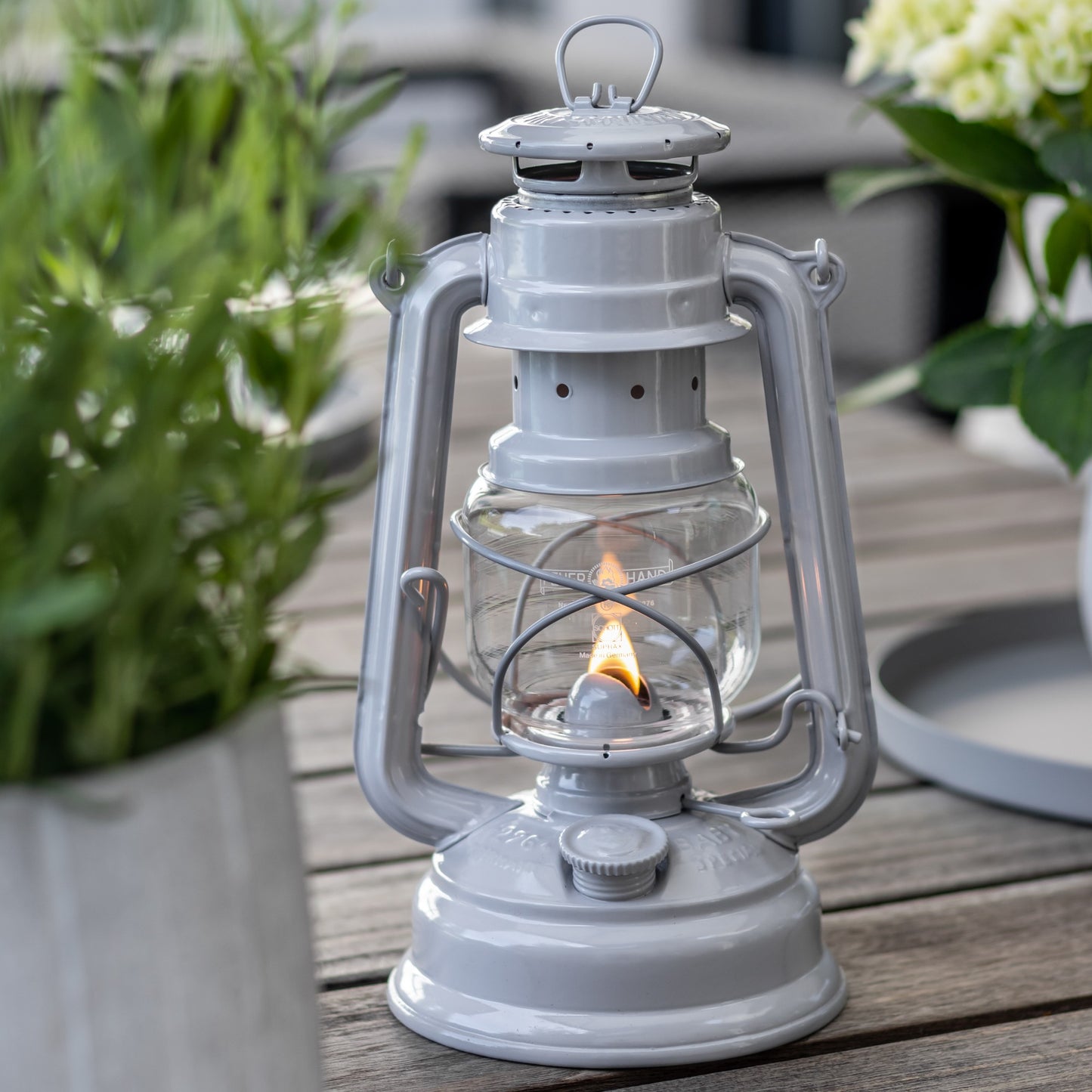 Handcrafted galvanized oil lantern with thermal heat-resistant globe, glowing softly on a wooden table surrounded by greenery, ideal for Lamps & Lighting, Camping, and Emergency Supplies collections.