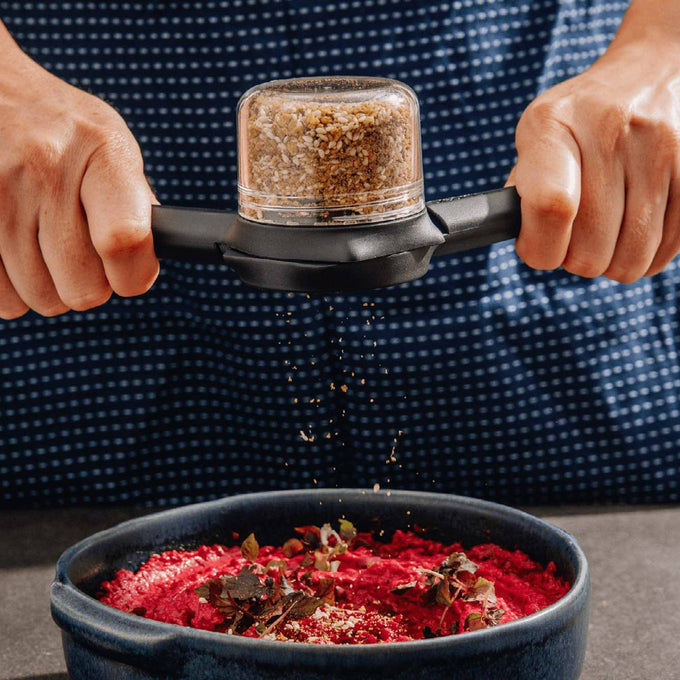 Hands squeezing black Ortwo Lite salt and pepper mill grinding coarse spice over a blue bowl of vibrant red dish, featured in Kitchen & Food and Kitchen Gadgets collections.