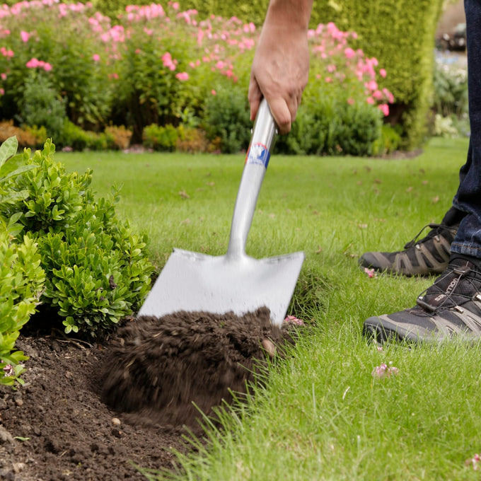 Person using a heavy-duty digging spade to turn soil in a garden bed next to green shrubs and manicured lawn, ideal for Farm, Garden & Tool, Digging and Cultivating, Lawncare, and Best Sellers collections.