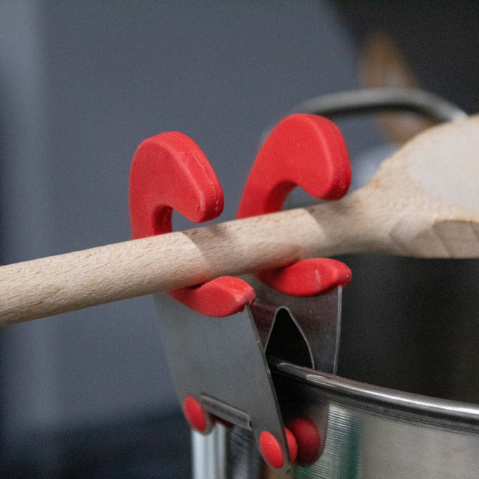Stainless steel pot clip with red silicone grips holding a wooden spoon securely off the pot rim, designed for Kitchen & Food, Kitchen Gadgets, and Cooking and Baking Helpers collections.