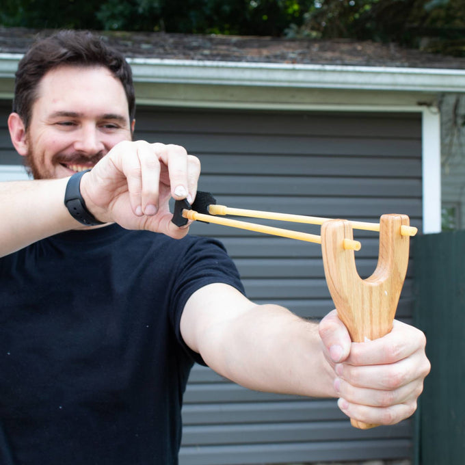Man outdoors aiming a handcrafted Amish made slingshot with smooth oak handle, black leather pouch, and yellow rubber tubing, featured in Toys, USA Made, and Lawn Games collections.