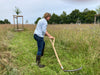 Person using a traditional curved blade scythe to cut tall grass in a rustic open field, showcasing practical farm and garden tool use for lawncare and home maintenance.