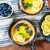Two handcrafted cast iron skillets with lemon and blueberry Dutch baby pancakes garnished with fresh mint, alongside a bowl of blueberries and sliced lemons, styled on a striped cloth for Kitchen & Food, Cast Iron, and USA Made collections.