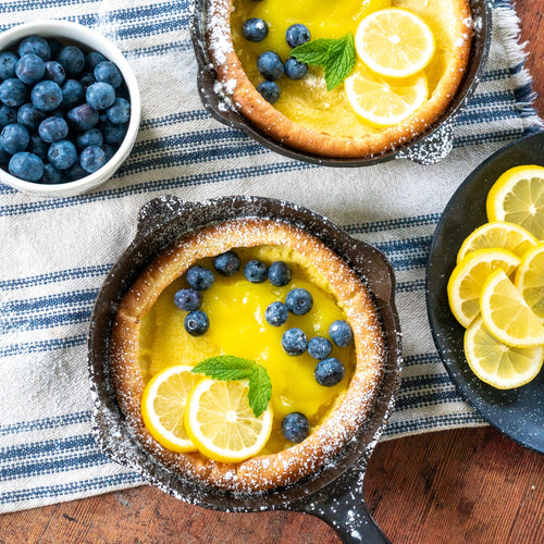 Two handcrafted cast iron skillets with lemon and blueberry Dutch baby pancakes garnished with fresh mint, alongside a bowl of blueberries and sliced lemons, styled on a striped cloth for Kitchen & Food, Cast Iron, and USA Made collections.