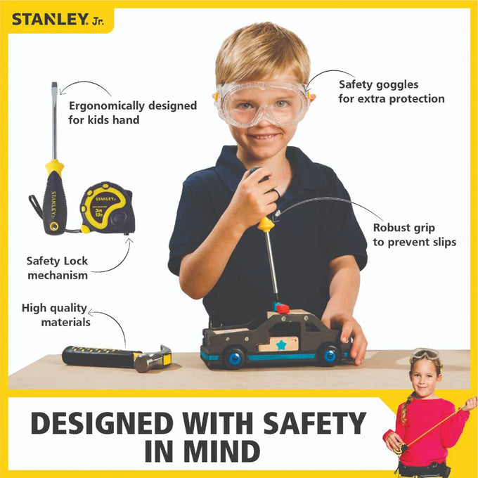 Young child wearing clear safety goggles and using a screwdriver on a wooden toy car, shown with additional hand tools featuring ergonomic yellow and black handles for indoor play and learning.