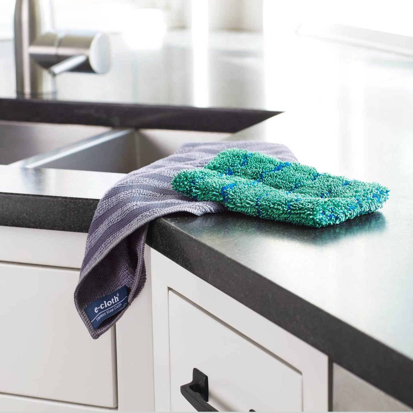 Green and blue textured microfiber cleaning cloth resting on a dark countertop with a folded gray microfiber cloth hanging over the edge, ideal for Home & Decor, Household Cleaning, and Best Sellers collections.