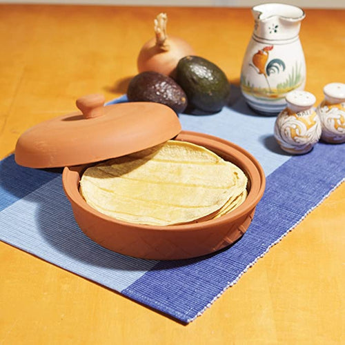 Handcrafted terra cotta tortilla warmer with lid slightly lifted, holding warm tortillas on a blue woven placemat, displayed alongside avocado, onion, and ceramic tableware, featured in Kitchen & Food, Storage and Serving, Best Sellers, and Promo Eligible collections.