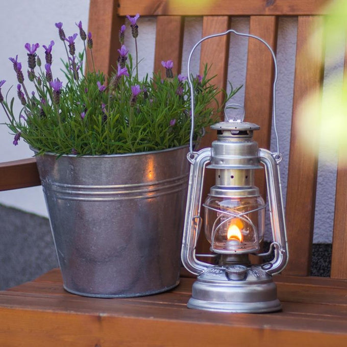 Handcrafted galvanized oil lantern with thermal heat-resistant globe glowing softly on a wooden bench beside a metal pot of purple flowers, perfect for Lamps & Lighting, Camping, and Emergency Supplies collections.