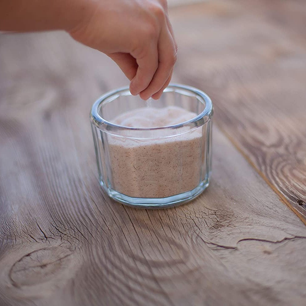 Hand sprinkling finely textured natural sea salt from a clear glass container on a rustic wooden surface, highlighting its pure, additive-free quality for Kitchen & Food, Pantry, USA Made, Best Sellers.