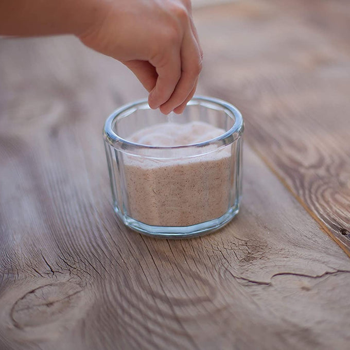 Hand sprinkling finely textured natural sea salt from a clear glass container on a rustic wooden surface, highlighting its pure, additive-free quality for Kitchen & Food, Pantry, USA Made, Best Sellers.