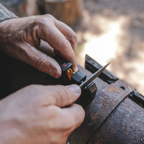 Hands sharpening a knife with a compact Work Sharp Pivot Plus, featuring ergonomic black and yellow components, set on a rustic metal surface, ideal for farm, kitchen, and hand tool use.