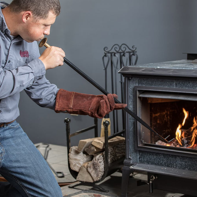 Person wearing a protective glove using a black powder-coated steel fire building utensil with brass mouthpiece to blow air into a wood stove fire, featured in Hearth Accessories and Stoves collections.
