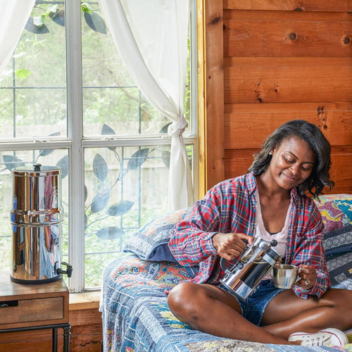 Woman sitting on bed in a rustic room pouring water from a polished stainless steel water filtration system with black elements, surrounded by cozy textiles, featured in homesteading and appliances collections.