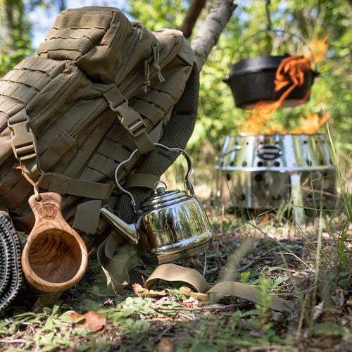 Polished stainless steel teakettle with bent handle hanging from a tan hiking backpack in a forest setting, near a portable silver stove with a black pot over an open flame, ideal for Farm, Garden & Tool, Kitchen & Food, Camping, Brewing Coffee and Tea, Best Sellers, and Promo Eligible collections.