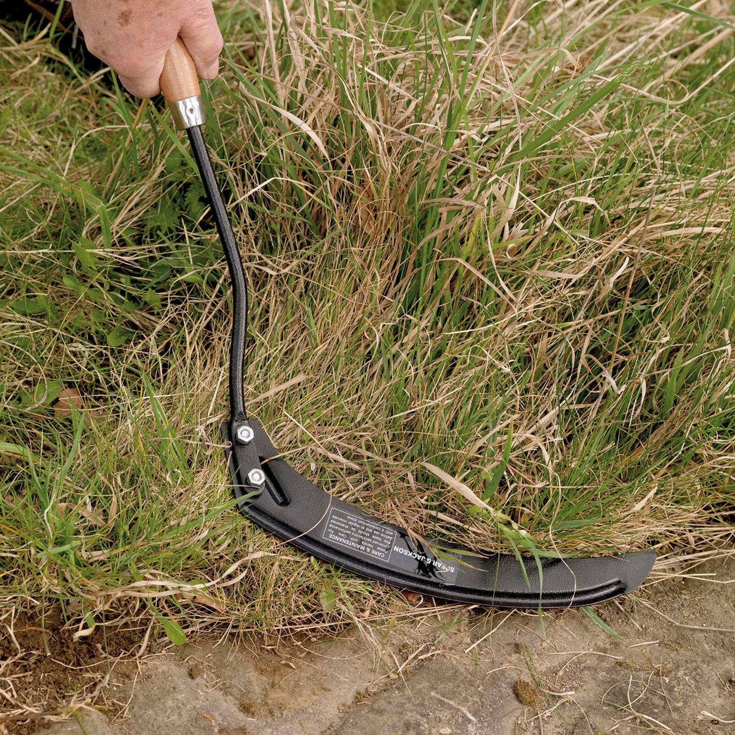 Close-up of a carbon steel grass hook with black hammer powder-coated blade and weatherproofed hardwood handle cutting tall grass, featured in Farm, Garden & Tool and Lawncare collections.